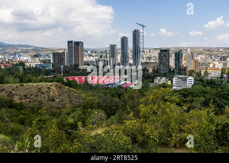 Vista sulla città di Tbilisi, Georgia. Sono visibili moderni edifici a torre e uno stadio con posti a sedere rossi. Una gru da costruzione si trova su uno degli edifici. La scena si trova nel quartiere Vake. Colline verdi circondano lo sviluppo urbano Foto Stock