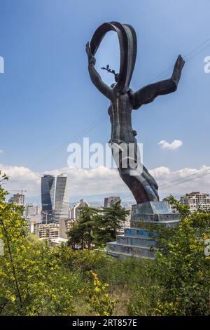 La statua in bronzo di una donna con le braccia alzate, che tiene uno stendardo e un ramo, corona il Memoriale della Gloria nel Vake Park a Tbilisi, Georgia. Lo stendardo oscilla in un arco sopra la sua testa. Sotto di lei, il plinto a gradini ricoperto di graffiti si trova tra arbusti gialli in fiore. A sinistra si ergono le torri Axis alte 147 metri su Ilia Chavchavadze Avenue, completate nel 2019. Aperto nel 1981, il memoriale con la Tomba del Soldato Sconosciuto commemora i soldati georgiani che hanno servito e sono morti nell'Armata Rossa durante la Seconda Guerra Mondiale. Lo scultore Giorgi Ochiauri ha creato le sue sculture. Il Vake Park è stato progettato a partire da Foto Stock