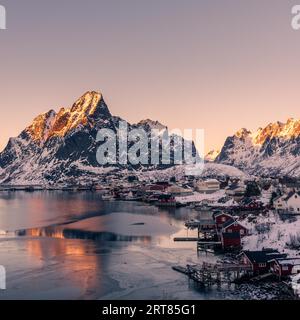 Il piccolo villaggio di pescatori Reine sulle isole Lofoten in Norvegia in inverno con ripide montagne innevate e lago ghiacciato durante il tramonto Foto Stock