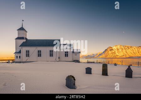 La piccola chiesa bianca in legno a Gimsoy sulla spiaggia sulle isole Lofoten in Norvegia in inverno, con un bellissimo cimitero vecchio e montagne in serata Foto Stock