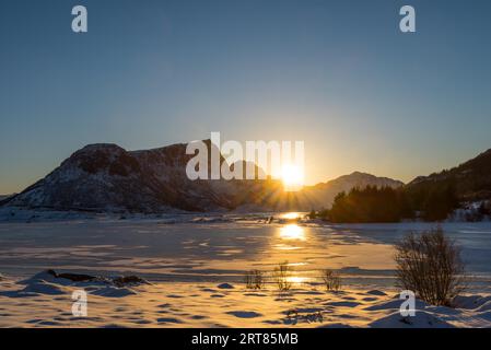 Sole ambientazione dietro montagne ricoperte di neve su lago ghiacciato in sogno Paesaggio invernale sulle isole Lofoten in Norvegia in chiaro giorno invernale Foto Stock