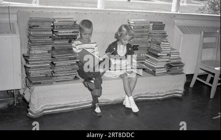 Anni '1950, storico, un giovane ragazzo e una ragazza seduti insieme a una panchina tra pile di libri rilegati, leggendo in un corridoio di una scuola elementare, Stati Uniti. Uno dei libri in cima a una pila è "Mr Popper's Penguins", pubblicato per la prima volta nel 1938 e nel 2011 trasformato in un popolare film per bambini. Foto Stock