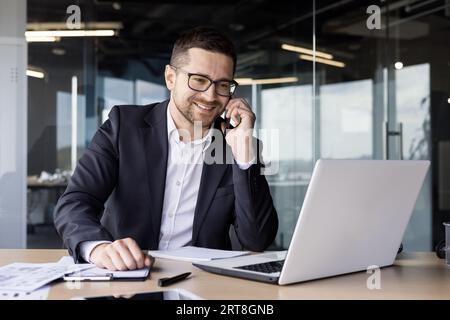 Un giovane avvocato sorridente, un notaio lavora in ufficio al portatile, parla e si consulta al telefono con i clienti. Foto Stock