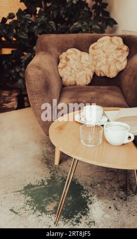 Tazze di caffè e libro su un tavolo di legno. Poltrona con cuscini e tavolo con tazze di caffè nel caffè. Tavolo per la colazione nel ristorante. Stile di vita urbano moderno Foto Stock