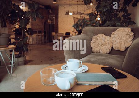 Tazze di caffè e libro su un tavolo di legno. Poltrona con cuscini e tavolo con tazze di caffè nel caffè. Tavolo per la colazione nel ristorante. Stile di vita urbano moderno Foto Stock