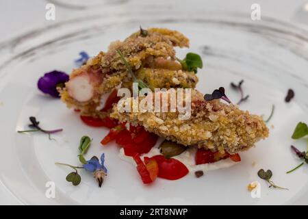 Piatto con polpo croccante impanato in panko su crema di stracciatella, pomodori datterini e olive Taggiasca. Fiori collocati intorno al cibo. Foto Stock