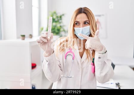 Giovane medico caucasico donna che tiene il test covid sorridente felice che punta con mano e dito Foto Stock