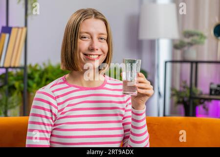 Donna sete di brunette che tiene il vetro di acqua naturale fare sorsi bere ancora acqua impedendo disidratazione si siede nel salotto di casa. Ragazza con buone abitudini di vita, sano dimagrimento, concetto di perdita di peso Foto Stock