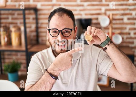 Uomo ispanico di taglia Plus con barba con moneta virtuale bitcoin sorridendo felice di puntare con mano e dito Foto Stock