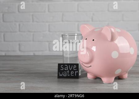 Scheda con frase Salva acqua. Salvadanaio e bicchiere di bevanda su tavolo di legno, spazio per testo Foto Stock