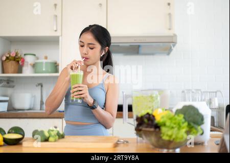 Una giovane donna asiatica bellissima e snella in abiti da palestra sta bevendo il suo frullato verde sano in cucina dopo il suo allenamento mattutino nel suo appartamento. Foto Stock