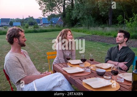 Tre amici europei si riuniscono intorno a un tavolo in un giardino sereno mentre il sole tramonta. Con un bicchiere di vino in mano, si impegnano in una conversazione calda e rilassata, assaporando la bellezza della serata e il legame di amicizia. Chiacchierate serali con gli amici europei in Garden at Sunset. Foto di alta qualità Foto Stock