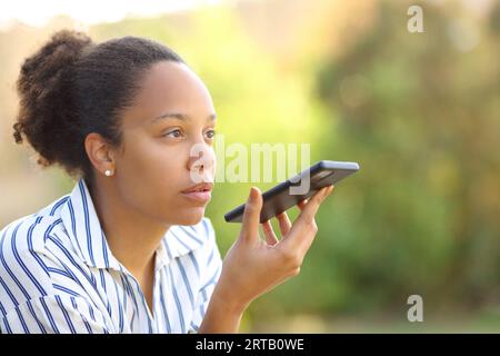 Donna di colore che detta testo sul telefono usig riconoscimento vocale seduto in giardino Foto Stock