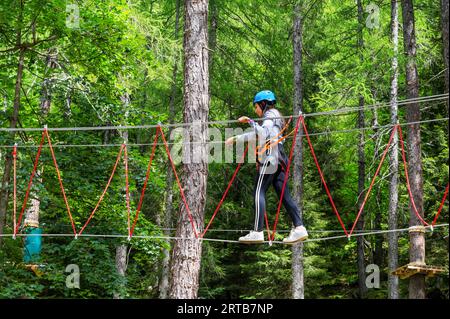 Vista laterale di una giovane donna con il casco che guarda verso il basso mentre cammina sulla corda sospesa e con cavi di sicurezza collegati nella foresta verde durante il giorno Foto Stock