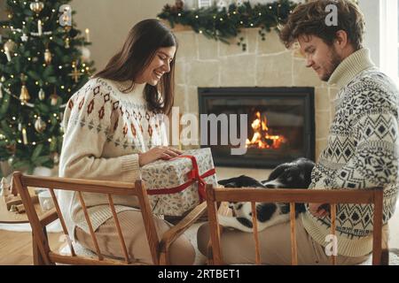 Felice coppia con intimi maglioni che giocano con il gatto e scambiano eleganti regali di natale sullo sfondo del camino con il mantello festivo e il cristo moderno Foto Stock