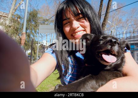 Ritratto selfie di una giovane donna latina seduta con il suo cane sorridente guardando la macchina fotografica, all'aperto nel parco al tramonto. Foto Stock