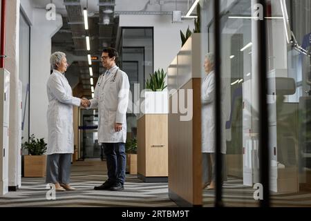 Due medici interculturali maturi in camici da laboratorio che stringono la mano e si guardano l'un l'altro mentre si trovano nel corridoio dell'ospedale e stanno facendo un accordo Foto Stock
