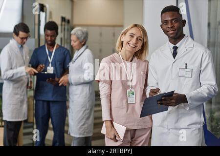 Due giovani medici interculturali di successo in uniforme che guardano la telecamera mentre si trovano contro un gruppo di colleghi che discutono di documenti Foto Stock