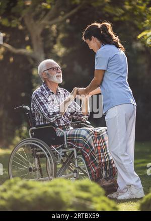 Giovane infermiera che tiene per mano un uomo anziano in sedia a rotelle nel parco Foto Stock