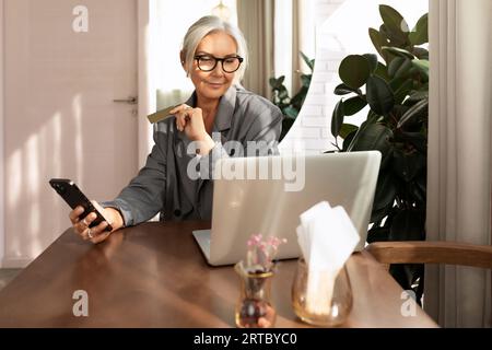 un'affascinante donna d'affari matura paga un ordine online utilizzando uno smartphone mentre si sta seduti in un bar e guarda uno smartphone Foto Stock