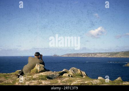 1954, storico, un gentiluomo che indossa una giacca sportiva e pantaloni seduti sulla cima di una scogliera erbosa a Lands End, affacciato sull'oceano con un binocolo, Cornovaglia, Inghilterra, Regno Unito. Foto Stock