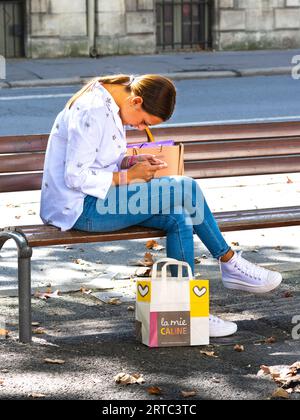 Giovane donna vestita in modo casuale seduta sulla panchina del centro città, studiando smartphone - Tours, Indre-et-Loire (37), Francia. Foto Stock