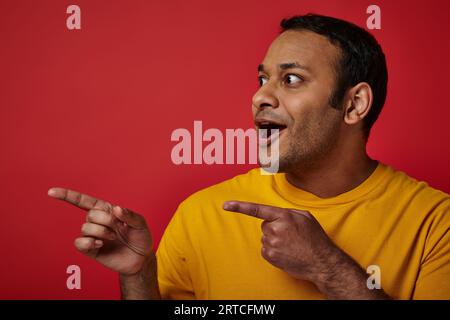 stupendo indiano con una t-shirt gialla che punta con le dita e guarda lontano sullo sfondo rosso Foto Stock