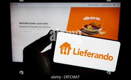 Persona che possiede uno smartphone con il logo della società tedesca di distribuzione di alimenti Lieferando sullo schermo di fronte al sito Web. Concentrarsi sul display del telefono. Foto Stock