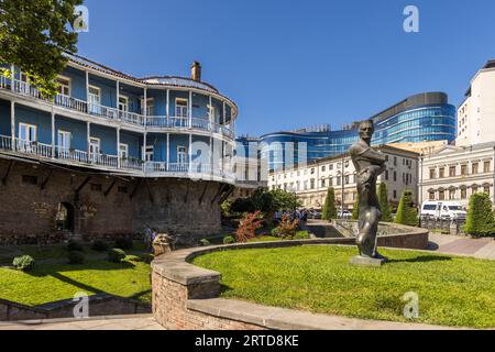 Una casa rotonda con balconi in legno dipinti di blu si trova nel viale Nikoloz Baratashvili nella vecchia città di Tbilisi, Georgia. La sua base in mattoni con archi rotondi è forse un residuo della vecchia muraglia cittadina. In primo piano, una slanciata scultura in bronzo di un uomo con le braccia incrociate si erge su un prato. In sfondo si innalza un edificio curvo con una facciata in vetro blu, accanto a case cittadine più antiche di colore pallido. Foto Stock