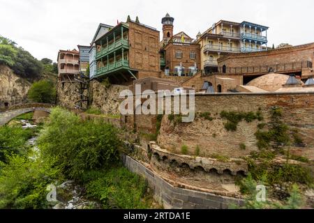 Edifici storici e bagni di zolfo caratterizzano il quartiere Abanotubani. Un fiume scorre attraverso il quartiere. Un ponte attraversa il fiume. La scena si trova a Tbilisi, Georgia Foto Stock