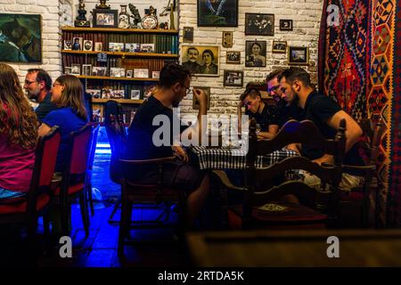 Gli ospiti sono seduti a tavoli con tovaglie a scacchi nella sala da pranzo del ristorante Nikolozi al 34 di via Anton Katalikosi nella vecchia città di Tbilisi, Georgia. Fotografie di famiglia incorniciate e quadri sono appesi alla parete di mattoni dipinta di bianco, e vecchi libri e foto sono disposti sugli scaffali. Un tappeto tessuto con motivi a rombi rossi, gialli e blu è appeso a destra. Una luce blu brilla in sottofondo. I proprietari Dodo e Beso gestiscono il ristorante. Scrivono il menu a mano, servono vino dalle loro stesse vigne e accettano solo contanti. Dodo è stato un calciatore professionista, per un periodo in Germania. Foto Stock