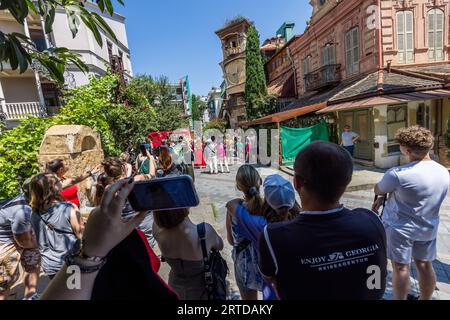 Gli spettatori aspettano davanti alla torre dell'orologio inclinata del Teatro delle Marionette Rezo Gabriadze al numero 13 di via Ioane Shavteli nella Città Vecchia di Tbilisi, Georgia, per lo spettacolo di mezzogiorno. In primo piano una mano tiene in alto uno smartphone. Un uomo indossa una maglietta con la scritta "Enjoy Georgia Reiseagentur". In inglese: "Enjoy Georgia travel agency". A destra si trova una casa con persiane in legno, un balcone in ferro battuto e un panno verde davanti all'ingresso. A sinistra, un blocco di pietra scolpito con una nicchia ad arco si erge davanti agli arbusti. Alle 12:00 e alle 19:00 un piccolo teatro delle marionette nella torre Foto Stock