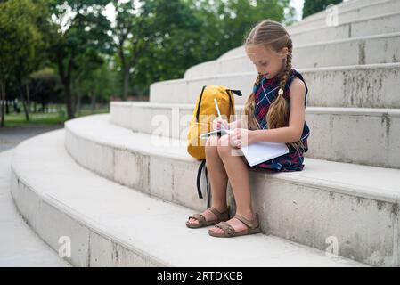 Un'adorabile studentessa, zaino al suo fianco, scrive nel suo notebook mentre si siede su gradini di pietra Foto Stock