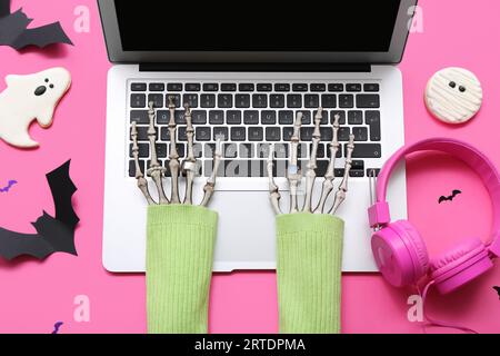Mani scheletriche con laptop, cuffie e decorazioni di Halloween su sfondo rosa Foto Stock