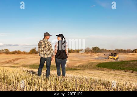 Vista da dietro di un marito e una moglie in piedi in un campo tenendosi per mano e trascorrendo un po' di tempo insieme mentre guardavano una mietitrebbia... Foto Stock