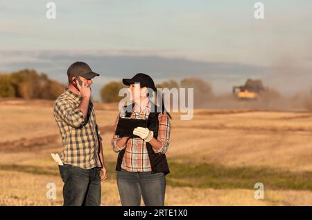 Marito e moglie in piedi in un campo che utilizzano dispositivi wireless portatili per gestire e monitorare la resa durante la raccolta di canola in autunno con un co... Foto Stock