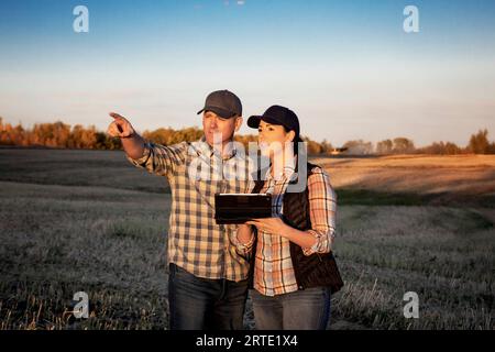 Un marito e una moglie in piedi nei campi al crepuscolo che utilizzano un dispositivo wireless portatile per gestire e monitorare la resa durante la loro caduta, canola ha... Foto Stock