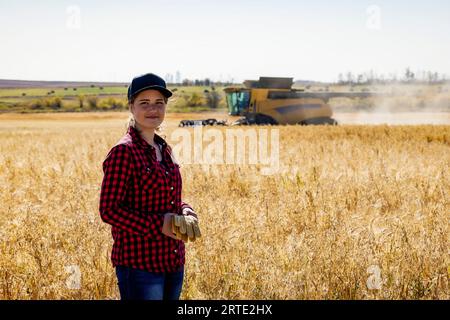 Ritratto di una giovane donna agricola in piedi in un campo di grano misto durante il raccolto, mentre una mietitrebbia lavora sullo sfondo Foto Stock