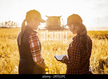 Vista ravvicinata da dietro di una donna agricola matura in piedi in un campo che lavora insieme a una giovane donna al momento del raccolto, usando agri avanzati... Foto Stock