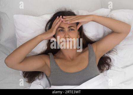 Una donna, i templi che lanciano, massaggia la testa, afferra il mal di testa, riposa in un letto soleggiato Foto Stock