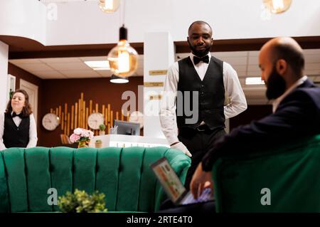 Fattorino che assiste un uomo d'affari nella lobby, fornisce servizi eccellenti mentre aspetta che la camera sia pronta per il check-in. Imprenditore che perde tempo con il laptop, scrivendo presentazioni per riunioni importanti. Foto Stock