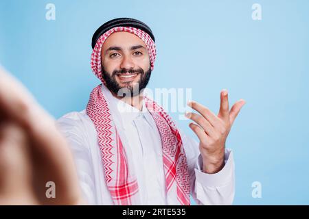 Allegro uomo musulmano che indossa abiti tradizionali che effettua videochiamate dal punto di vista pov. Persona araba sorridente in stoffa e sciarpa che guarda la fotocamera anteriore mentre parla in una riunione online Foto Stock