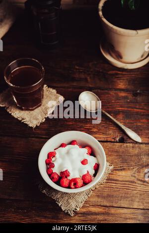 Ciotola di dessert salutare con lamponi maturi e yogurt e bicchiere di caffè su superficie di legno Foto Stock