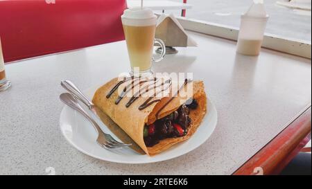 Vista orizzontale di una crepe di frutta rossa con cioccolato, contenente fragole, more, uva e cioccolato fuso, con un cappuccino in t Foto Stock