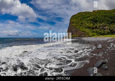 Vista panoramica dell'Oceano Pacifico e delle scogliere ricoperte di alberi sulla riva di una spiaggia di sabbia lavica nera con surf e cielo nuvoloso blu lungo la strada ... Foto Stock