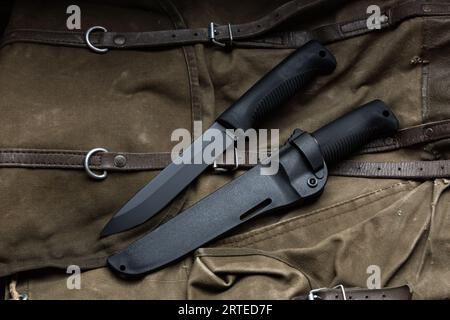 Coltello moderno con manico in gomma e guaina in plastica. L'acciaio freddo si trova su uno zaino da trekking vintage. Sfondo di legno. Foto Stock