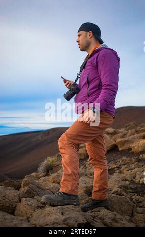 Uomo in piedi su rocce vulcaniche sulla cima di una montagna sopra le nuvole di Haleakala guardando l'alba sulla costa del Pacifico con uno smartphone wi... Foto Stock