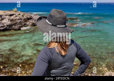 Vista ravvicinata da dietro di una donna che indossa un cappello da sole in piedi sulla riva guardando le acque turchesi sul fronte oceano di Kaanapali Bea... Foto Stock