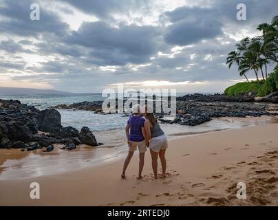 Vista presa da dietro di una madre matura con la figlia adulta che si tiene per mano e si trova sulla sabbia della spiaggia segreta guardando il co... Foto Stock