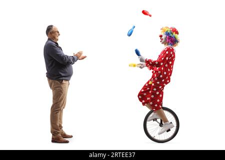 Foto a profilo completo di un uomo maturo che applaude un clown che cavalca un uniciclo e giocola isolato su sfondo bianco Foto Stock